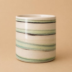 Peut inclure: Un gobelet cylindrique en céramique présentant une base de couleur crème avec des rayures horizontales peintes à la main dans des tons vert sauge et brun foncé. Le récipient a une finition lisse et brillante avec un profil droit.