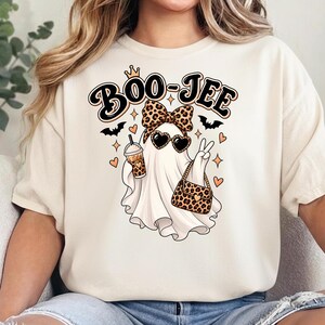 May include: A cream-colored t-shirt featuring a graphic of a ghost wearing a leopard print bow and heart-shaped sunglasses. The ghost holds a leopard print cup and handbag, making a peace sign. The text BOO-JEE is printed above in black.