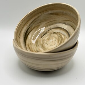 May include: Two stacked ceramic bowls featuring a swirling, marbled pattern in shades of beige, cream, and dark brown. The bowls have a smooth, glossy finish with visible horizontal ridges along the exterior walls and a deep, rounded interior basin.