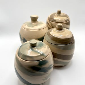 May include: A set of four round ceramic jars with matching lids, each featuring a unique marbled pattern in shades of cream, sage green, and dark brown. The containers have a smooth, glossy finish and small, knob-shaped handles on the lids.