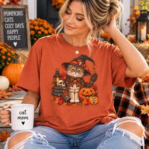 May include: A burnt orange t-shirt featuring a graphic of a cat wearing a witch hat, surrounded by a lantern, a skull, stacked books labeled Spells, Treats, and Cats, autumn leaves, and a jack-o'-lantern. A white mug with the text cat mom is held.