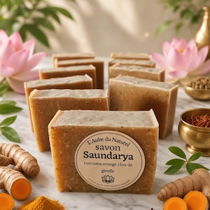 Puede incluir: Pastillas de jabón rectangulares de color tostado con motas visibles, cada una con una etiqueta redonda beige que dice L'Aube du Naturel savon Saundarya curcuma, orange clou de girofle, dispuestas en filas con raíces de cúrcuma y clavos.