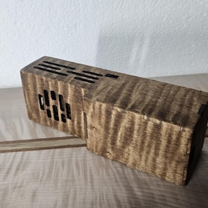 Peut inclure: Une boîte rectangulaire en bois avec un motif de grain ondulé et une finition brun foncé. Les panneaux supérieurs et latéraux présentent des fentes de ventilation découpées au laser disposées en grille. La boîte est assemblée par tenons.