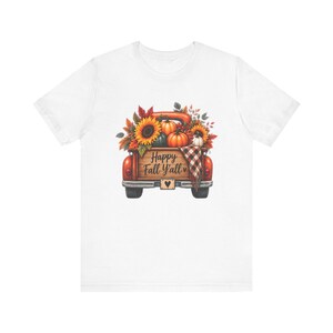 May include: A white t-shirt featuring a graphic of a red pickup truck bed filled with sunflowers, orange and white pumpkins, and autumn leaves. The truck bed has the text Happy Fall Y'all printed on it, along with a plaid blanket and a heart icon.