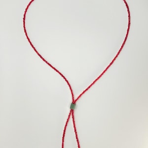 May include: A necklace composed of small, vibrant red beads strung on a thin cord. The necklace features a single, muted green cylindrical bead that acts as a sliding toggle, allowing the two ends of the red beaded cord to hang down freely.