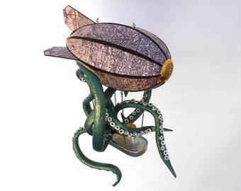 May include: A whimsical steampunk-style airship sculpture. The airship features a map-patterned body with black accents and a brass-colored front. It is supported by teal-colored octopus tentacles, creating a unique and imaginative design.