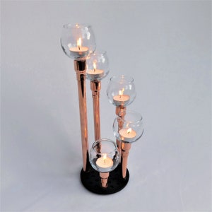 Könnte beinhalten: Ein kupferner Kerzenhalter mit fünf gläsernen Kerzenhaltern. Die Kerzenhalter sind in einer kaskadierenden Anordnung angeordnet. Der Kerzenhalter steht auf einem weißen Hintergrund.