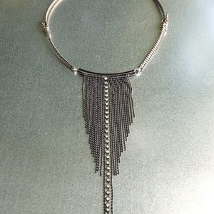 Könnte beinhalten: Eine silberfarbene Halskette mit Fransenanhänger. Die Halskette hat einen Kettenkragen und einen zentralen Anhänger mit mehreren Strängen kleiner, dunkler Perlen. Eine Reihe klarer Kristalle verläuft in der Mitte der Fransen.