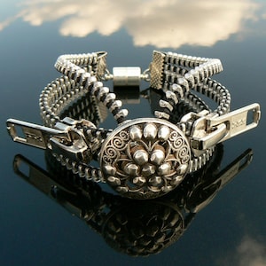 May include: A silver zipper bracelet with a decorative button closure. The bracelet is made of three layers of zipper teeth and has a silver metal clasp.