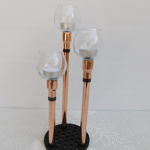 Könnte beinhalten: Ein kupferner Kerzenhalter mit drei gläsernen Kerzenhaltern. Der Kerzenhalter hat eine schwarze Basis mit einem kreisförmigen Muster.