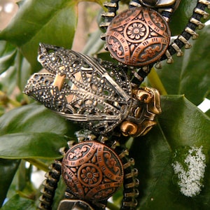 May include: A close-up of a decorative zipper charm featuring a detailed, intricate design of a cicada with a copper-toned finish. The charm is attached to a black zipper with copper-toned accents.
