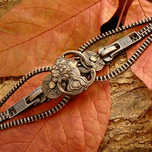 May include: A zipper bracelet with a decorative metal charm featuring a woman's face. The charm is in a gold tone and has intricate details. The bracelet is made of black and silver zippers.