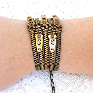 May include: A unique bracelet made from multiple black and gold zippers. The bracelet features gold zipper pulls and a black chain closure. The zippers are arranged side by side, creating a bold and edgy accessory.