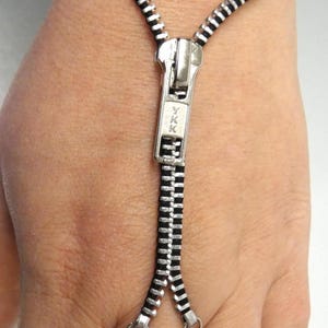 May include: A silver-coloured zip bracelet with a black backing. The zip pull is visible, and the bracelet extends to a finger ring. The bracelet is designed to be worn on the wrist and connected to the finger.