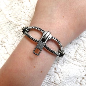 May include: A silver-toned bracelet designed to resemble a zipper. The bracelet is wrapped around a wrist, with the zipper pull in the open position. The bracelet's teeth are detailed and the pull has a rectangular shape.