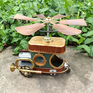 May include: A steampunk-style sculpture of a vehicle with a copper propeller, gold accents, and a brass horn. The body is a mix of teal, gold, and copper, with decorative circular frames. The sculpture is set against a backdrop of green foliage.