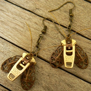 May include: A pair of gold-toned earrings featuring a decorative butterfly design with a zipper pull in the center. The zipper pull has the text "YKK" on it.