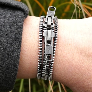 May include: A silver-coloured zipper bracelet with three rows of zipper teeth. The bracelet is fastened with a zipper pull, and the YKK logo is visible. The bracelet is worn on a wrist, with a dark grey sleeve in the background.