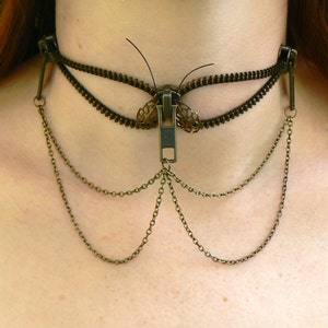May include: A gold chain choker necklace with a zipper pull in the shape of a butterfly. The zipper pull is made of metal and has a black finish. The chain is made of multiple strands and has a delicate design.