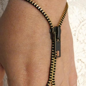 Puede incluir: Una pulsera única con diseño de cremallera. La pulsera está hecha de dientes de metal dorado con un respaldo negro y un tirador de cremallera a juego. Se lleva en la muñeca y está conectada a un anillo.