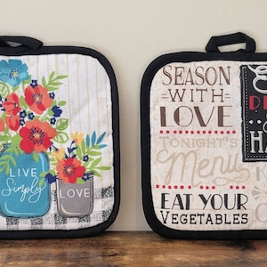 May include: Two white cotton pot holders with black trim. One pot holder features a floral design with three mason jars that say "Hope", "Live Simply", and "Love". The other pot holder has a chalkboard design with the text "Season with Love", "Tonight's Menu", "Eat Your Vegetables", "Eat, Drink & be Happy", "Kiss the Cook".