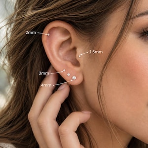 Puede incluir: Un conjunto de seis pendientes pequeños redondos con piedras claras, con tamaños etiquetados de 1,5 mm, 2 mm, 3 mm y 4 mm. Los pendientes están dispuestos en varios piercings en el lóbulo, el hélix y el trago, mostrando diferentes diámetros.