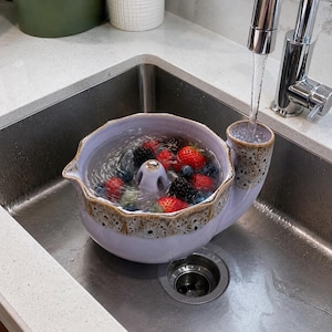 Könnte beinhalten: Ein hellviolettes Beeren-Sieb aus Keramik mit gewelltem Rand und gesprenkelter, braun texturierter Kante. Die Schale verfügt über einen zentralen Abflusskegel und ist mit Wasser sowie frischen Erdbeeren, Blaubeeren und Brombeeren gefüllt.