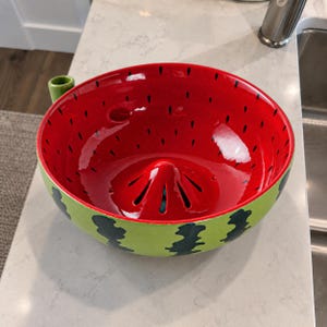 Peut inclure: Un presse-agrume en céramique en forme de pastèque. L'intérieur est rouge vif avec des motifs de pépins noirs et un cône central, tandis que l'extérieur est vert clair avec des rayures verticales vert foncé et une petite poignée verte.