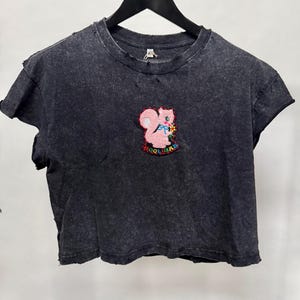 Op de afbeelding: Een antracietgrijs acid-wash T-shirt met een ronde hals en gerafelde randen. In het midden bevindt zich een geborduurde patch van een roze eekhoorn die een gele bloem vasthoudt, met het woord HOOLIGAN in veelkleurige letters eronder.