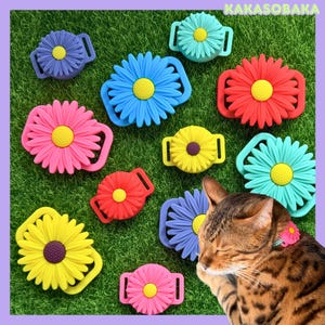 May include: A collection of silicone daisy-shaped charms in various colors including blue, red, pink, yellow, and purple, each featuring a central yellow or dark brown disc. One charm is attached to a collar worn by a brown tabby cat.