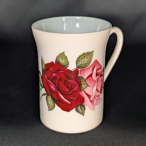 May include: A cream-coloured ceramic mug featuring a floral design with one large deep red rose and one smaller light pink rose, accompanied by green leaves. The mug has a smooth, cylindrical body and a curved handle on the right side.