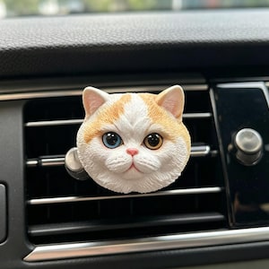 Peut inclure: Un clip désodorisant pour voiture en forme de tête de chat avec une fourrure blanche et orange. Le chat a un œil bleu et un œil ambre, un petit nez rose et une apparence texturée et réaliste fixée sur une grille d'aération noire.