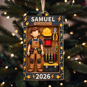 Puede incluir: Un adorno colgante rectangular con el nombre SAMUEL y el texto Electrician y 2026. Muestra una figura con peto marrón y cinturón de herramientas, casco amarillo, cable, alicates, destornilladores y una caja de herramientas negra y amarilla.