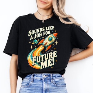 May include: A black short-sleeve t-shirt featuring a graphic of a rocket ship with a colorful exhaust trail, a small planet, and stars. The text reads "SOUNDS LIKE A JOB FOR FUTURE ME!" in a bold, retro-style font with yellow and white lettering.