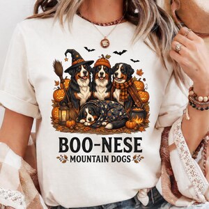 May include: A cream-colored t-shirt featuring a graphic of four Bernese Mountain Dogs in Halloween attire, including a witch hat and scarf, surrounded by pumpkins, bats, and a lantern, with the text BOO-NESE MOUNTAIN DOGS printed below.