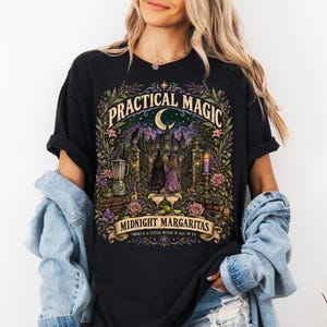 May include: A black t-shirt featuring a colourful graphic print with the text "PRACTICAL MAGIC" and "MIDNIGHT MARGARITAS" along with "THERE'S A LITTLE WITCH IN ALL OF US". The illustration depicts a castle, two figures, a blender, and margaritas.