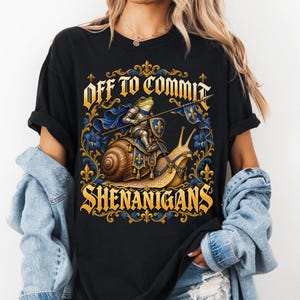 May include: A black t-shirt featuring a graphic of a frog knight in silver armour riding a large snail. The frog holds a spear and a blue shield with a gold fleur-de-lis. The text reads "OFF TO COMMIT SHENANIGANS" in gold lettering.