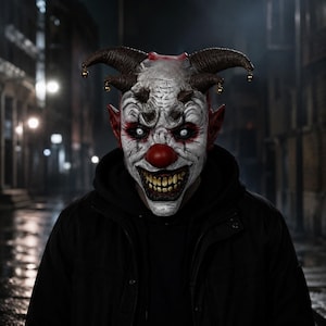 Puede incluir: Una máscara de payaso de cabeza completa con un rostro blanco texturizado, una nariz roja brillante y maquillaje de ojos rojo oscuro. La máscara tiene dos grandes cuernos curvos con pequeñas campanas doradas, orejas rojas y dientes amarillos.