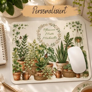 May include: A rectangular mouse pad featuring a botanical illustration of various potted plants, including a snake plant and monstera, with a central wreath enclosing the text "Pflanzen Mutti Michaella". A white computer mouse rests on the surface.