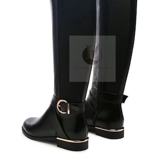 May include: A pair of black knee-high boots featuring a smooth finish, a side zip, and a decorative strap at the ankle with a gold-toned buckle. The heel and sole edge are accented with a gold-toned metallic trim. A logo reads LEATHER LEGACY.