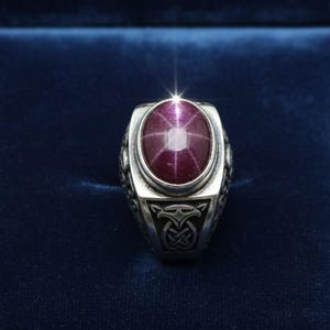 May include: A silver ring featuring a large, oval-shaped cabochon star ruby with a distinct six-rayed star effect. The metal band displays intricate, dark-oxidized Celtic-inspired knotwork and bird-like engravings on the sides of the setting.