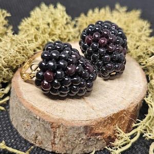 May include: A pair of earrings shaped like blackberries, featuring small, glossy, dark purple and black spherical beads clustered together to mimic the texture of real fruit, attached to simple gold-toned metal earring findings.