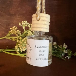 May include: A small clear glass bottle car diffuser with a light-colored wooden cap and a hanging rope cord. The white label on the front features black text that reads Rosemary Mint Car Diffuser, with additional partial text visible on the side.
