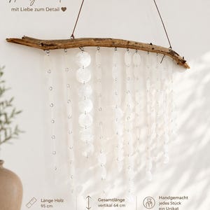 Op de afbeelding: Een wandhanger met een horizontaal stuk drijfhout van 95 cm lang en 3 cm dik. Meerdere strengen van doorschijnende, ronde witte schelpschijfjes hangen aan het hout. De totale verticale lengte is 64 cm.