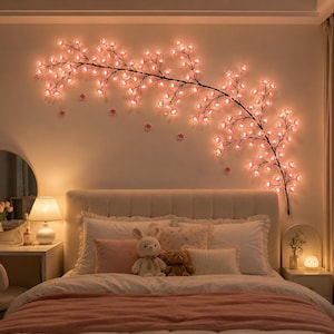 Op de afbeelding: Een decoratieve wandlamp in de vorm van een uitgestrekte kersenbloesemtak met donkere stelen en talloze kleine, gloeiende roze bloemvormige ledlampjes, horizontaal geplaatst op de muur boven een bed.