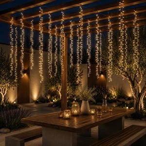 Op de afbeelding: Een reeks verticale lichtsnoeren met warmwitte ledlampen die aan een houten pergola boven een rechthoekige houten eettafel hangen. De tafel is voorzien van diverse glazen kaarsenhouders en een centrale witte vaas met groen.