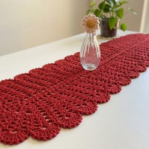 May include: A long, rectangular, deep red crochet table runner featuring a repeating scalloped edge pattern. The runner is laid out flat and has a detailed, open-work stitch design throughout its entire length, showcasing a consistent, intricate texture.