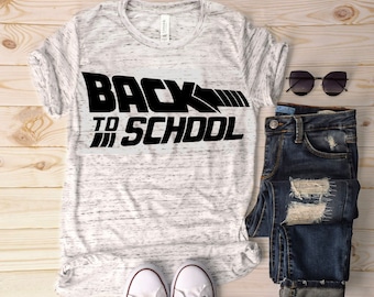 May include: A white t-shirt with a black graphic that says "Back to School" in a stylized font. The shirt is paired with blue jeans, white sneakers, and sunglasses.