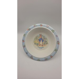 May include: A round white ceramic bowl featuring a central illustration of Peter Rabbit in a blue jacket standing in a garden. The rim is decorated with a light blue border patterned with small colorful vegetables and garden motifs.