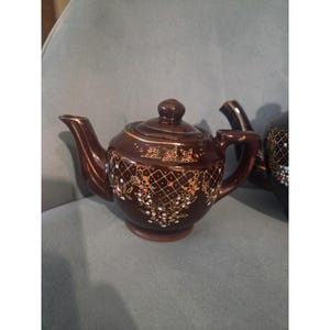 May include: A dark brown ceramic teapot with a matching lid, featuring a gold-colored lattice pattern and small floral accents in orange and white. The teapot has a curved spout and a handle, with a glossy finish reflecting light on its surface.